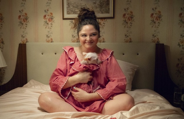 Megan Stalter as Jessica in Too Much holding a dog and sat cross legged on her bed wearing pink pyjamas