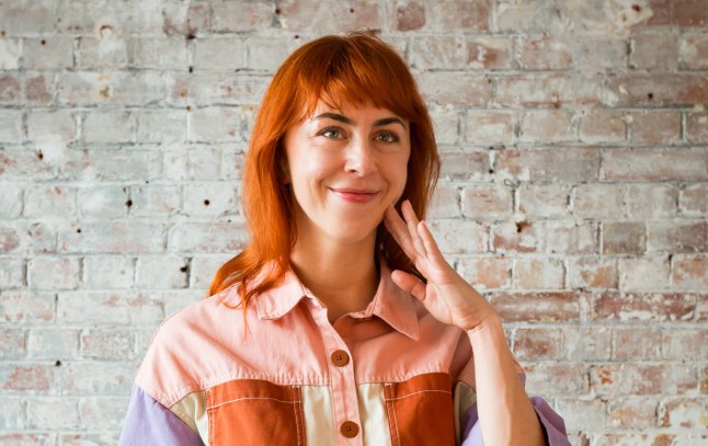 Candace Bryan with hand on face, red hair, and pastel coloured shirt.